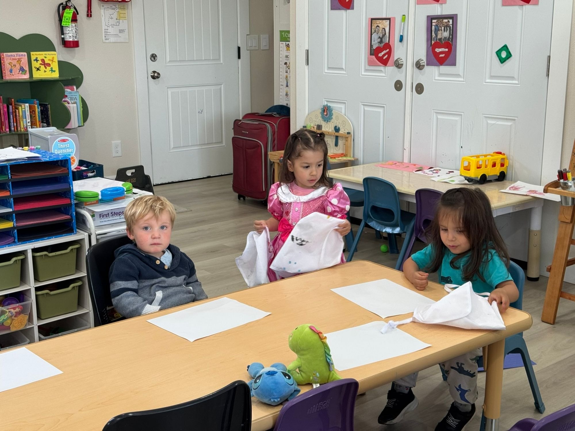 Three children putting on smocks at the table before an art activity