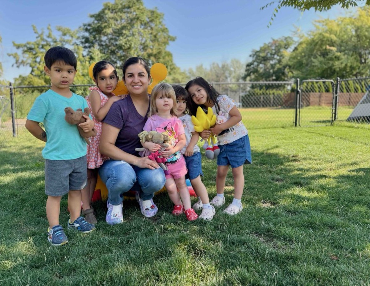 A teacher sitting on the grass with five smiling children holding stuffed toys