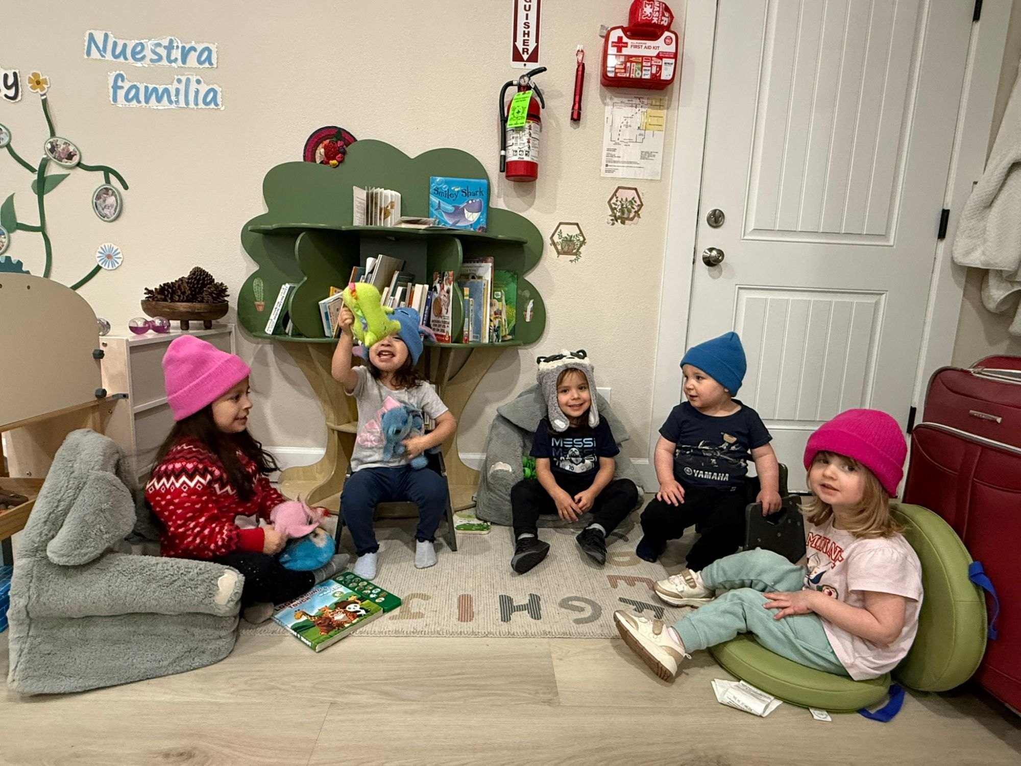 Five children in knit hats sitting on soft chairs around a tree-shaped bookshelf