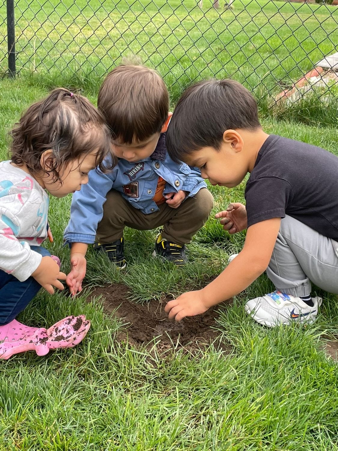 Three children crouched on the grass, digging in a patch of dirt
