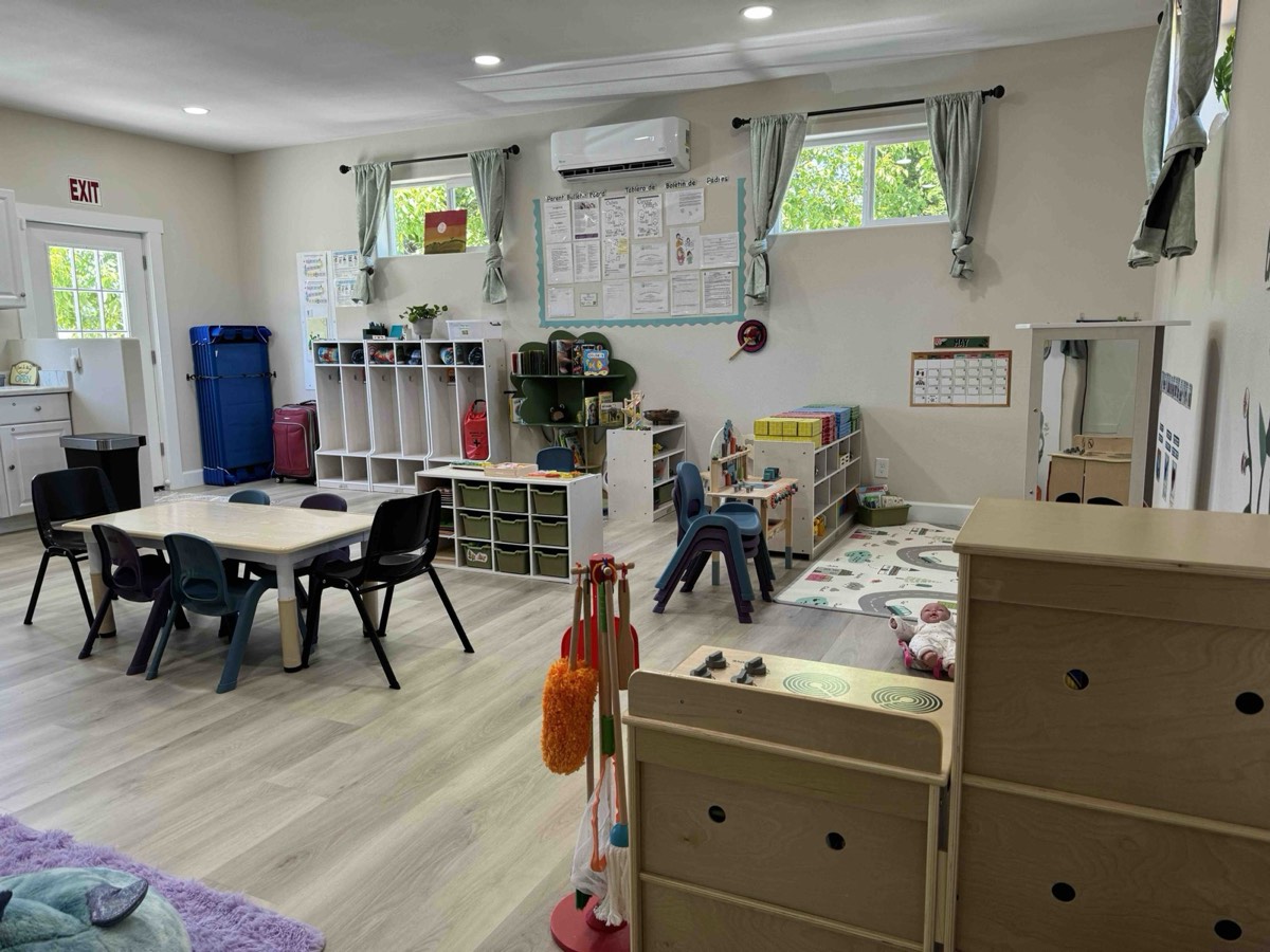 A bright playroom with small tables, shelves of toys and a play rug