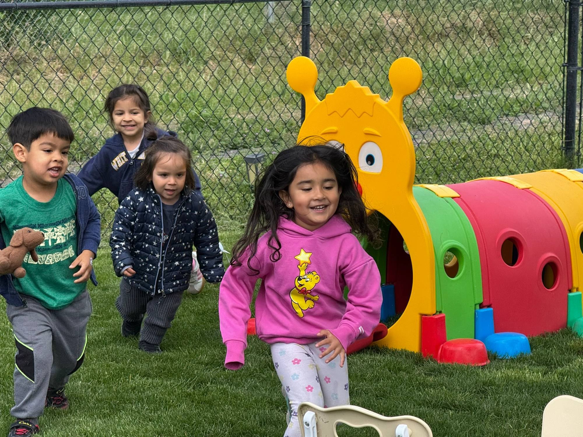 Four smiling children running across the grass past a colorful caterpillar tunnel