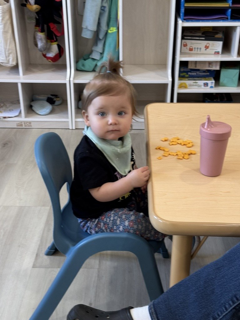 A toddler in a bib sitting at a small table with a snack and a sippy cup