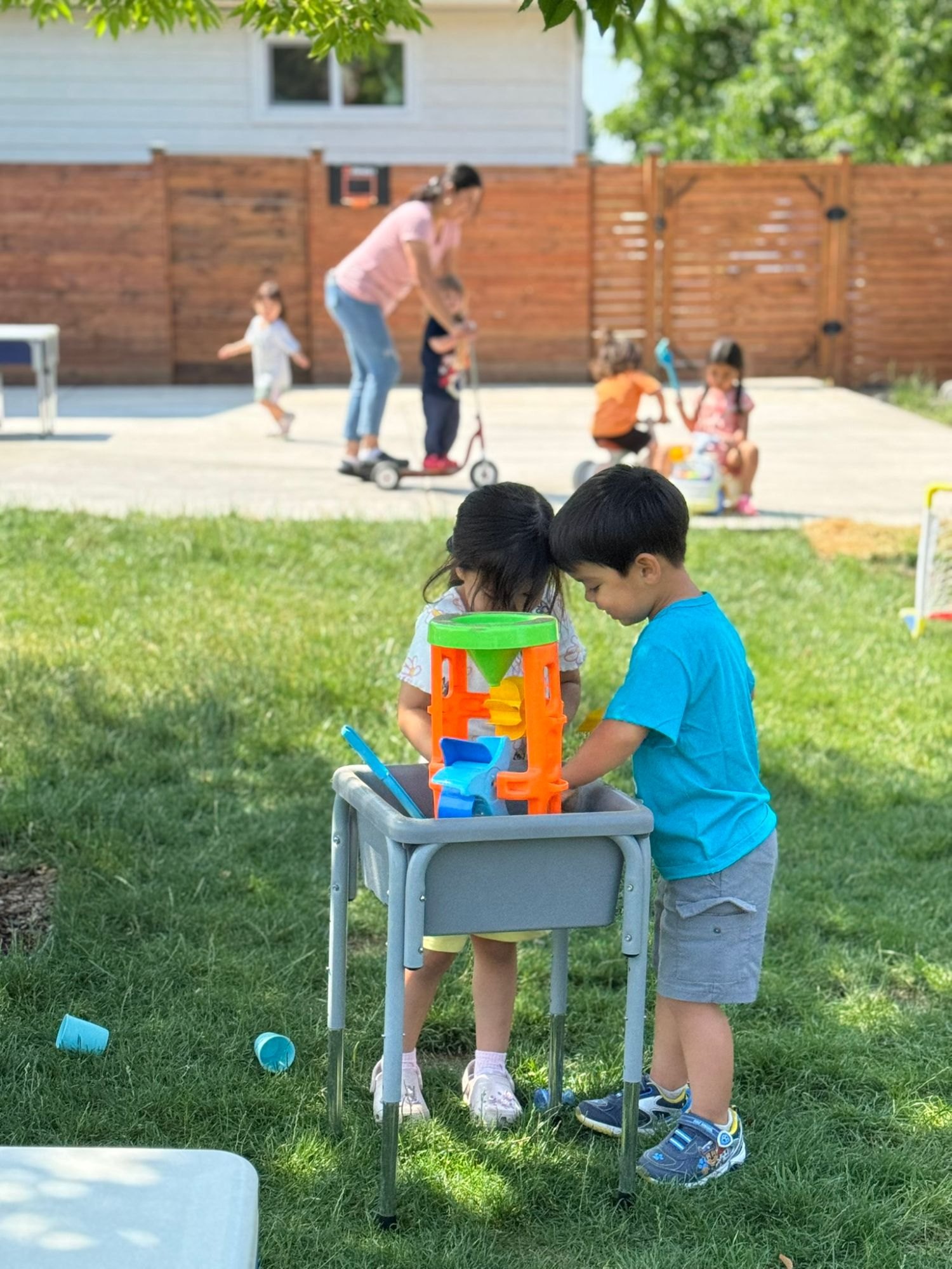 Two children playing at a water table with a water wheel, while Antonia helps a child on a scooter behind them